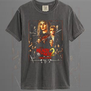 May include: A pepper-colored short-sleeve t-shirt featuring a graphic print of the movie poster for Ready or Not 2. The design includes multiple character portraits, a red and yellow title logo, and the text Here I Come on a dark background.