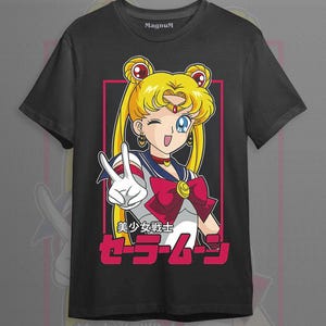 May include: A black short-sleeve t-shirt featuring a graphic of the anime character Sailor Moon with blonde hair in buns, wearing a sailor-style uniform with a red bow, making a peace sign. The text includes Japanese characters and Sailor Moon.