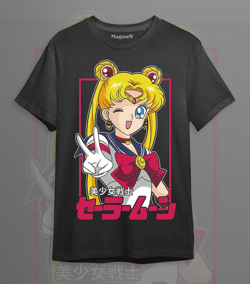 May include: A black short-sleeve t-shirt featuring a graphic of the anime character Sailor Moon with blonde hair in buns, wearing a sailor-style uniform with a red bow, making a peace sign. The text includes Japanese characters and Sailor Moon.