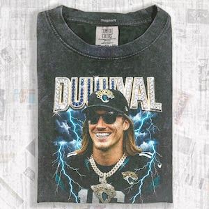 May include: Dark gray Comfort Colors t-shirt featuring a graphic of a smiling person wearing sunglasses and a baseball cap. The word "DUUUVAL" is displayed in a jeweled font. Blue lightning bolts are in the background.