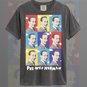 Può includere: T-shirt color pepe con una grafica in stile pop art di Pee-wee Herman. La grafica è composta da nove quadrati con la stessa immagine di Pee-wee Herman su sfondo rosso, giallo e blu. Il testo "PEE-WEE HERMAN" è in basso.