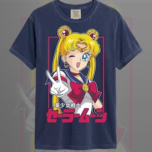 May include: A navy blue short-sleeve t-shirt featuring a graphic of a blonde character with twin buns making a peace sign. The design includes a red bow, a blue collar, and the Japanese text 美少女戦士セーラームーン in pink and white lettering.