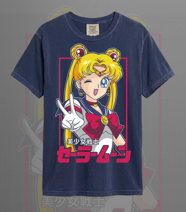 May include: A navy blue short-sleeve t-shirt featuring a graphic of a blonde character with twin buns making a peace sign. The design includes a red bow, a blue collar, and the Japanese text 美少女戦士セーラームーン in pink and white lettering.