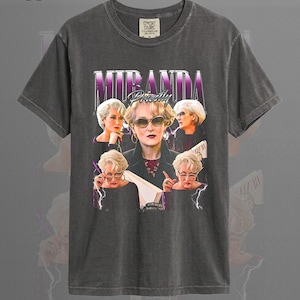 Puede incluir: Camiseta color pimienta con un gráfico de Miranda Priestly de la película "El diablo viste de Prada". El diseño incluye múltiples imágenes del personaje con el nombre "Miranda Priestly" en morado y blanco.