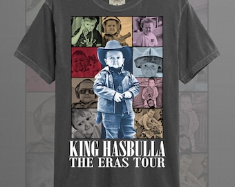 May include: Graphite-colored t-shirt featuring a collage of images and the text "KING HASBULLA THE ERAS TOUR." The central image is a child in a cowboy hat and jacket. The shirt is made of a soft, comfortable material.