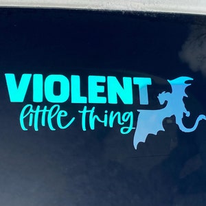 May include: A teal vinyl decal on a car window. The decal features the words "VIOLENT little thing" in a script font, with a dragon silhouette to the right. The background is a dark, reflective surface.