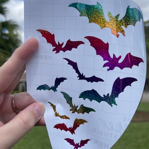 May include: A sheet of clear transfer paper with multiple iridescent rainbow glitter bats of varying sizes. The bats are arranged in a staggered pattern, with the largest bat at the top and the smallest bat at the bottom.