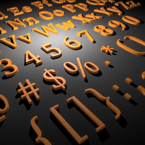 Può includere: Un assortimento di lettere, numeri e simboli 3D di colore arancione, tra cui il simbolo del dollaro, la percentuale e le parentesi, sono sparsi su una superficie grigio scuro. L'illuminazione crea ombre, dando agli elementi un aspetto dimensionale.