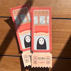 May include: Two red and white ticket-shaped bookmarks with a cartoon character from the movie Spirited Away. The character is a black and white spirit with a white face and a black body. The text on the bookmark reads "Spirited Away - One Ticket".