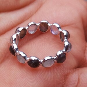 May include: A silver ring featuring a series of small, round, multi-coloured stones set around the band. The stones appear to be a mix of light blue, grey, and dark grey. The ring is displayed on a person's hand.