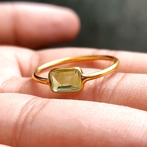 Op de afbeelding: Een gouden ring met een rechthoekige, lichtgroene edelsteen. De ring is gezet op een dunne gouden band. De ring wordt getoond op een open handpalm.