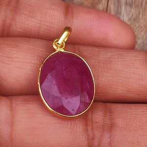 Peut inclure: Un pendentif en or avec une grosse pierre précieuse de rubis ovale et facettée. Le rubis est d'un rouge profond et est serti dans une lunette en or. Le pendentif est tenu dans une main sur un fond en bois.