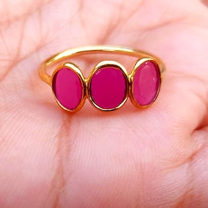 May include: A gold-toned ring featuring three horizontally aligned oval-shaped fuchsia pink stones set in a smooth metal band. The design showcases a continuous row of vibrant pink cabochons held within a polished gold-coloured metal setting.