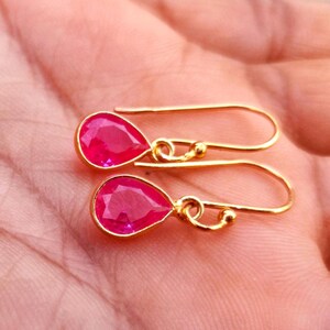 Könnte beinhalten: Ein Paar goldfarbene Ohrhänger mit facettierten, tropfenförmigen, leuchtend pinken Steinen. Jeder Ohrring hängt an einem einfachen goldfarbenen Haken, der eine kräftige, gesättigte pinke Farbe und eine polierte, reflektierende Oberfläche aufweist.