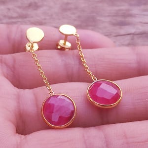 May include: A pair of gold-tone drop earrings featuring round, faceted, deep pink stones set in polished metal bezels. Each earring is suspended from a delicate gold-tone chain attached to a flat, circular stud post with a secure backing.