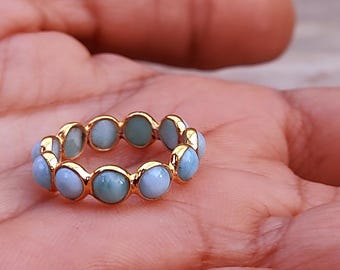 May include: A gold-colored ring featuring a series of light blue gemstones. The ring is set with multiple round, pale blue stones, creating a continuous band. The ring is displayed on a person's palm.