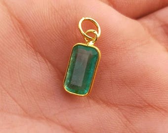 May include: A gold pendant featuring a rectangular emerald-cut green gemstone. The pendant has a small gold loop for a chain. The gemstone is a vivid green and set in a gold bezel. The pendant is likely for a necklace.