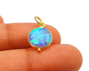 May include: A gold pendant with a round, iridescent opal stone. The opal displays shades of blue and green. The pendant has a small loop at the top for a chain and a decorative bead at the bottom. Jewelry.