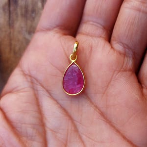 May include: A gold-coloured pendant featuring a teardrop-shaped, vivid red gemstone. The pendant hangs from a small gold loop. The jewellery is shown on an open palm.