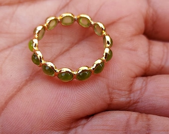 Könnte beinhalten: Ein goldener Ring mit einer Reihe ovaler, hellgrüner Edelsteine. Der Ring wird in einer Hand präsentiert, wobei das Design zur Geltung kommt. Das Goldband des Rings ergänzt die grünen Steine und erzeugt einen klassischen Look.