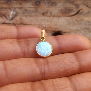 Könnte beinhalten: Goldener Anhänger mit einem runden, hellblauen Opalstein. Der Opal hat eine schimmernde, irisierende Qualität. Der Anhänger wird von einer kleinen Goldschlaufe gehalten. Der Hintergrund ist eine Holzoberfläche.