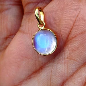 May include: A gold-colored pendant with a round, iridescent, blue and white stone. The pendant has a small loop at the top for a chain. The stone appears to be a moonstone. The pendant is held in a person's hand.
