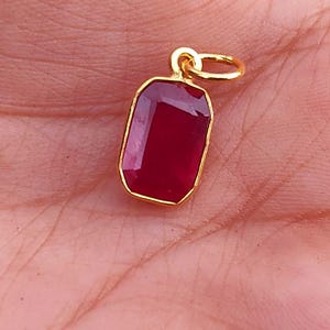 May include: A gold-colored pendant with a rectangular, deep red gemstone. The pendant has a simple, elegant design with a small loop at the top for a chain. The gemstone is set in a gold bezel.