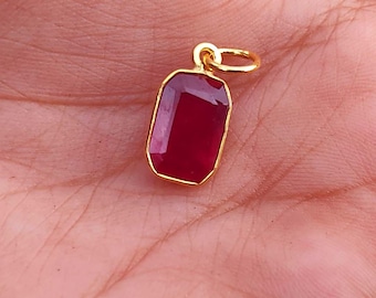 Op de afbeelding: Een goudkleurige hanger met een rechthoekige, dieprode edelsteen. De hanger heeft een eenvoudig, elegant ontwerp met een kleine lus aan de bovenkant voor een ketting. De edelsteen is gezet in een gouden zetting.