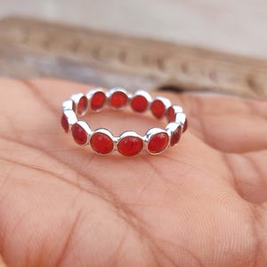 Può includere: Un anello d'argento con una serie di pietre preziose rosse ovali. Le pietre sono distanziate uniformemente attorno alla circonferenza dell'anello. L'anello è mostrato su uno sfondo neutro. Un gioiello.