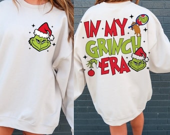 May include: Cream-coloured sweatshirt with a Grinch face in a Santa hat on the front. The back features the text "IN MY GRINCH ERA" in red and green, plus Grinch graphics and a Christmas ornament. A festive holiday apparel item.