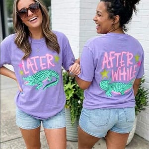 May include: Two women wearing matching lavender t-shirts with a cartoon alligator design. The front of the shirts say "LATER" and the back says "AFTER WHILE" in pink lettering with star accents. They are also wearing denim shorts.