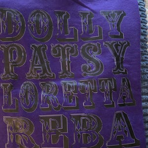May include: A purple t-shirt featuring the names DOLLY, PATSY, LORETTA, and REBA printed in a bold, black, decorative serif font stacked in four rows. The shirt is folded and placed on a textured blue surface with a white patterned shirt below.