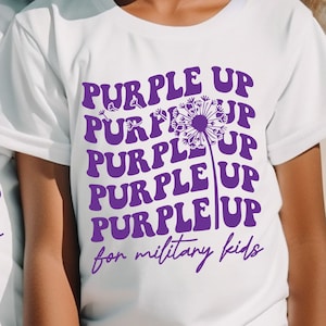 Puede incluir: Dos camisetas blancas con texto morado que dice "PURPLE UP" repetido cinco veces con un gráfico de diente de león morado. El texto "for military kids" está debajo del gráfico.