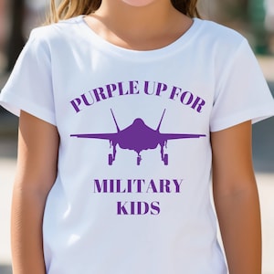 Puede incluir: Camiseta blanca con un diseño morado que dice "Purple Up For Military Kids" y presenta la silueta de un avión de combate.