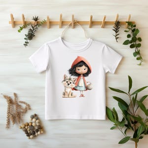 Könnte beinhalten: Weißes T-Shirt mit einer Cartoon-Illustration eines Mädchens in einem roten Kapuzenmantel und einem Wolf. Das Mädchen hält einen Korb. Der Wolf sitzt daneben. Das Design verwendet weiche Pastellfarben.