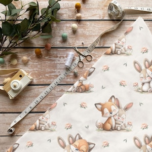 Könnte beinhalten: Draufsicht auf einen Stoff mit einem sich wiederholenden Muster aus Füchsen und Hasen, zusammen mit Nähwerkzeugen und dekorativen Elementen. Ein Maßband zeigt Zentimeter. Die Szene umfasst eine Schere, Faden und eine Spielzeugkamera, alles auf einer Holzoberfläche angeordnet.