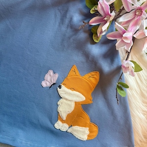 May include: A light blue skirt featuring an orange fox applique with white accents and a pink butterfly. The skirt has a soft, velvety texture. A dark blue fabric with woodland creatures is visible at the top.