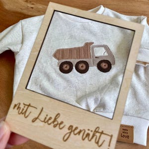 May include: A light-colored baby sweatshirt with a brown and tan dump truck applique. A wooden frame with the words "mit Liebe genäht!" is in front of the sweatshirt. A small tag that says "made with love" is on the sleeve.