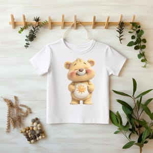 Könnte beinhalten: Weißes T-Shirt mit einem lächelnden Cartoon-Bären, hellgelb mit einem Sonnensymbol auf dem Bauch. Das T-Shirt hängt an einem Holzgestell. Ein fröhliches Design für Kinder, mit Grünpflanzen im Hintergrund.