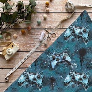 Könnte beinhalten: Ein teal- und blau gemusterter Stoff mit einem sich wiederholenden Design von Videospiel-Controllern. Die Controller haben einen Farbspritzer-Effekt. Ein Maßband, eine Schere und Faden sind ebenfalls sichtbar.