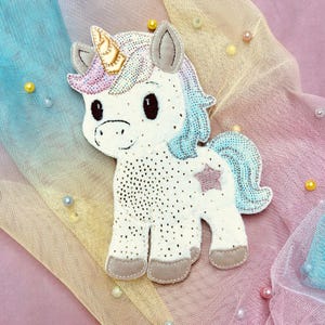 以下が含まれることがあります： 金色の角、ピンクの星、青とピンクのたてがみと尾を持つ白いユニコーン型のアップリケ。 アップリケは穴あきボディで、散らばった真珠のあるパステルレインボーの背景に配置されています。