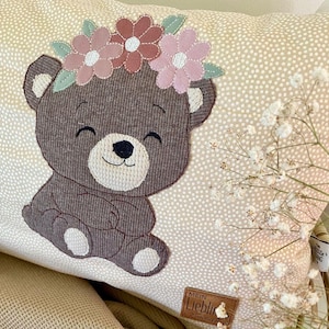 Könnte beinhalten: Ein dekoratives Kissen mit einem lächelnden Bären, verziert mit einem Blumenkranz. Der Bär ist braun mit cremefarbenen Details. Das Kissen hat einen cremefarbenen Hintergrund mit weißen Punkten. Ein kleines Lederetikett ist sichtbar.