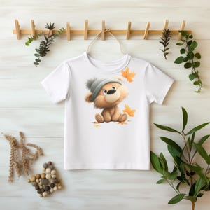 Könnte beinhalten: Weißes T-Shirt mit einem Cartoon-Bären, der eine graue Mütze trägt und von orangefarbenen Herbstblättern umgeben ist. Der Bär sitzt auf einem Blätterbett. Das T-Shirt hängt an einem Holzgestell.
