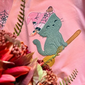 May include: Pink Halloween-themed sweatshirt featuring an embroidered design of a green cat riding a broomstick, wearing a witch hat. The witch hat is pink with a spiderweb design. The background is pink with spiderwebs.