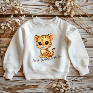 Könnte beinhalten: Weißes Sweatshirt mit einem Cartoon-Leoparden mit braunen Flecken und großen Augen. Der Text "Frech, Wild, Wunderbar" ist unter dem Leoparden aufgedruckt. Das Sweatshirt hat lange Ärmel und einen Rundhalsausschnitt.