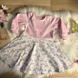 Könnte beinhalten: Ein rosa Kleid mit langen Ärmeln und einem weißen Rock mit Regenbogenmuster. Der Oberteil des Kleides hat einen rosa Hasen mit einer Schleife gestickt.