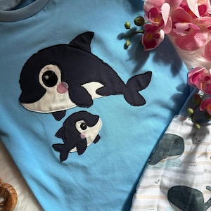 May include: A light blue baby outfit featuring a large and small orca whale applique. The orcas are navy blue with white accents and pink cheeks. The outfit includes a long-sleeved top and pants with a whale pattern.