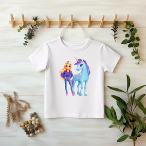 Könnte beinhalten: Weißes T-Shirt mit einer Cartoon-Illustration eines Mädchens und eines Einhorns. Das Mädchen hat blondes Haar, eine blaue Jacke und blaue Jeans. Das Einhorn ist blau mit einer rosa und lila Mähne und einem Horn. Das T-Shirt hängt an einem Holzgestell.