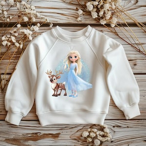 Może przedstawiać: Biała bluza z nadrukiem z kreskówkową ilustracją księżniczki w niebieskiej sukni i renifera. Projekt umieszczono na jasnoniebieskim tle. Bluza ma długie rękawy i okrągły dekolt.