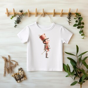 Może przedstawiać: Biała koszulka z kreskówkową ilustracją żołnierza w czerwonym i złotym mundurze, trzymającego pałeczkę. Żołnierz nosi wysoki, ozdobny kapelusz. Koszulka wisi na drewnianym wieszaku z dekoracyjną zielenią.